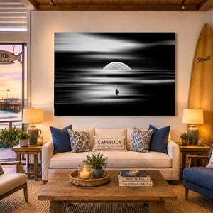 May include: A large rectangular wall art print featuring a black and white long exposure photograph of a lone figure standing on a beach under a large moon, with horizontal motion blur across the sky and water, displayed above a beige sofa.