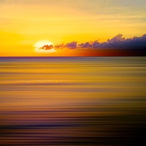 May include: A single bird stands in the shallows of a beach at sunset. The water is a blur of golden and purple hues, reflecting the colors of the sky. The sun is setting behind a line of dark clouds.