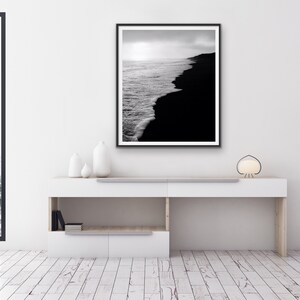 May include: Black and white photograph of a beach with waves crashing on the shore. The photo is framed in a black frame and hanging on a white wall.