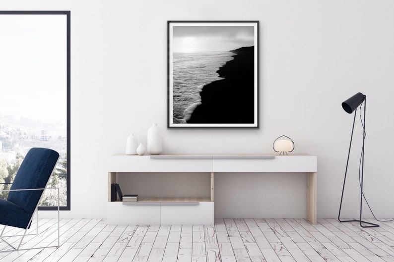 May include: Black and white photograph of a beach with waves crashing on the shore. The photo is framed in a black frame and hanging on a white wall.