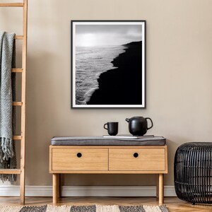 May include: Black and white photograph of a black sand beach with white waves crashing on the shore. The photo is framed in a black frame and hanging on a wall.
