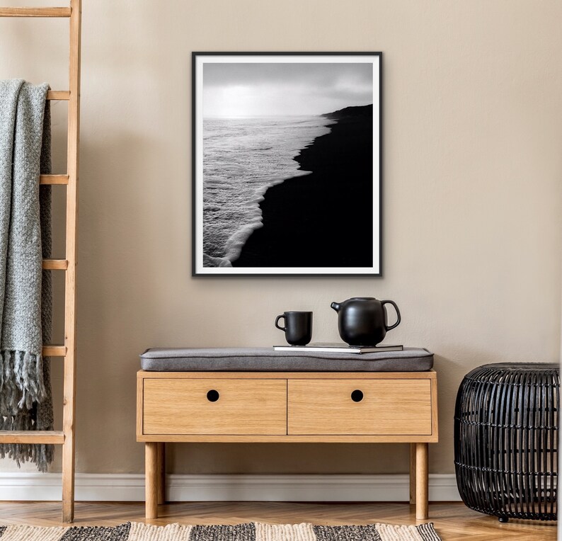 May include: Black and white photograph of a black sand beach with white waves crashing on the shore. The photo is framed in a black frame and hanging on a wall.