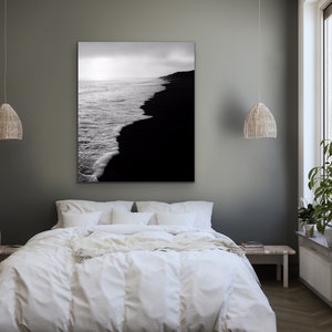May include: A black and white photograph of a beach with waves crashing on the shore. The photo is hanging on a gray wall above a white bed with white bedding.