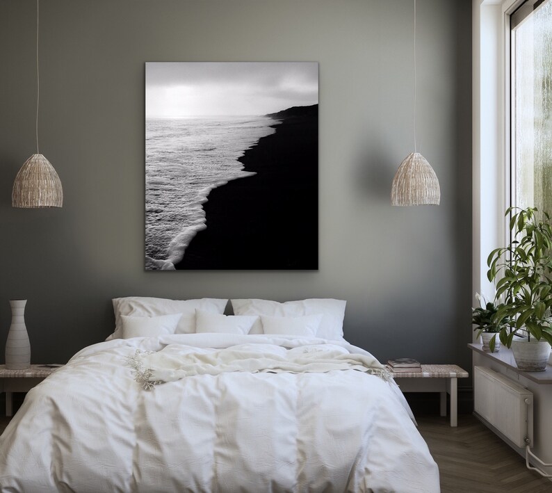 May include: A black and white photograph of a beach with waves crashing on the shore. The photo is hanging on a gray wall above a white bed with white bedding.