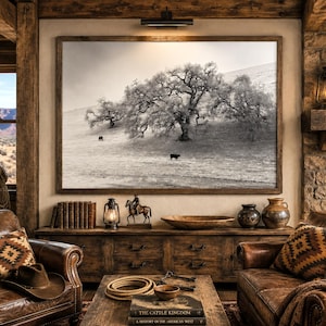 May include: A large framed black and white landscape photograph of a sprawling oak tree on a grassy hillside with two grazing cattle, mounted on a wall above a rustic wooden console table featuring a collection of books and decor items.