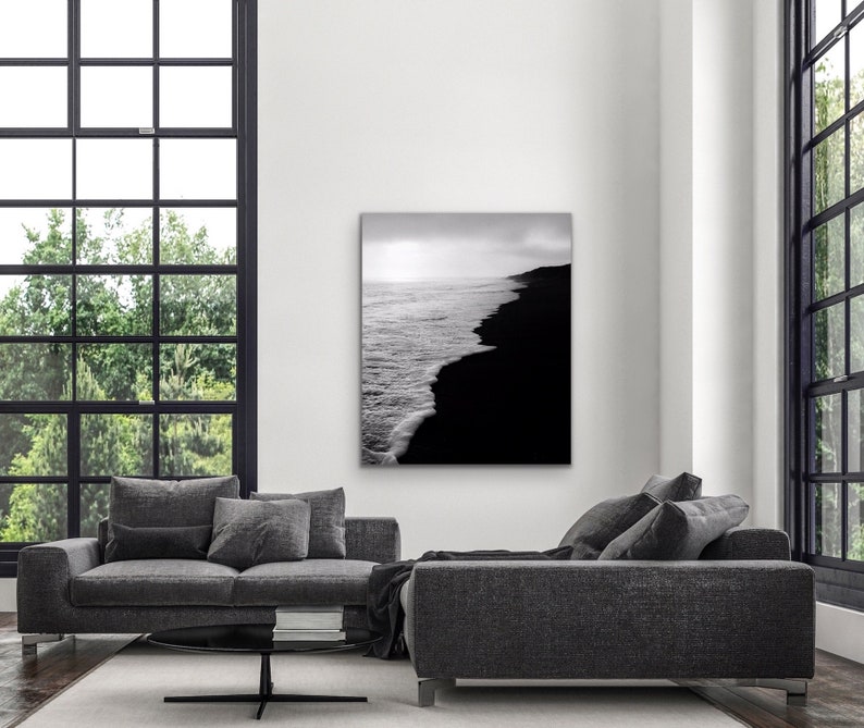 May include: Black and white photograph of a beach with waves crashing on the shore. The water is on the left side of the image and the sand is on the right side.