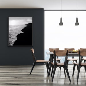 May include: A black and white photograph of a beach with waves crashing on the shore hangs on a black wall above a dining table with six wooden chairs. The chairs have black metal legs and are arranged around a wooden table with a gray tabletop.