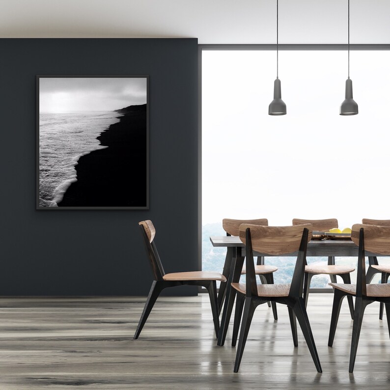 May include: A black and white photograph of a beach with waves crashing on the shore hangs on a black wall above a dining table with six wooden chairs. The chairs have black metal legs and are arranged around a wooden table with a gray tabletop.