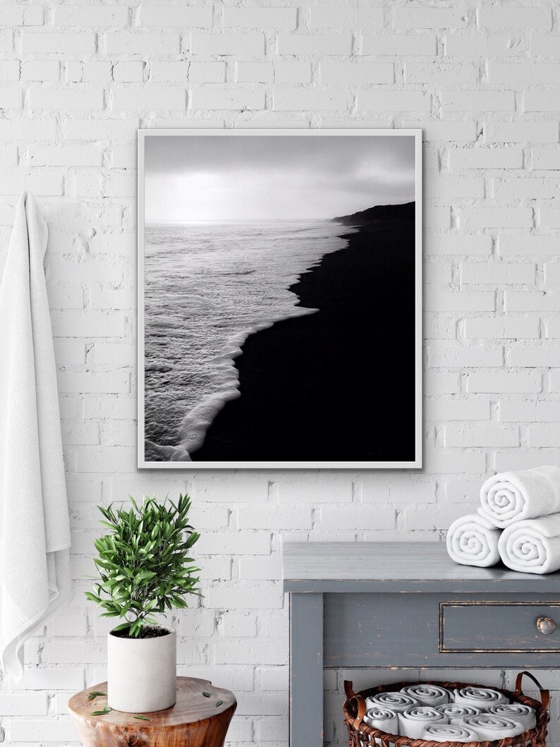 May include: Black and white photograph of a beach with waves crashing on the shore. The sand is black and the water is white. The photo is framed in a white frame.