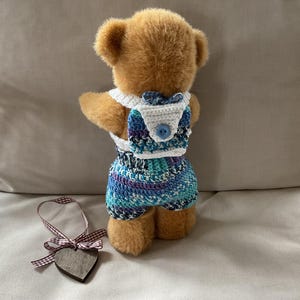 Könnte beinhalten: Ein brauner Teddybär, der ein blaues und weißes Häkel-Outfit mit einem weißen Häkel-Rucksack trägt. Der Rucksack hat einen blauen Knopf.