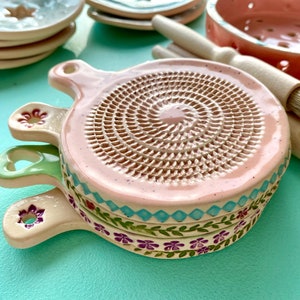 Puede incluir: Una pila de ralladores de cerámica hechos a mano en tonos rosa, verde y crema. Cada rallador tiene una superficie de rallado circular y un asa con un diseño floral decorativo. Otros utensilios de cocina de cerámica son visibles en el fondo.