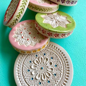 Könnte beinhalten: Eine Sammlung von fünf runden Keramikuntersetzern mit komplexen geprägten Mandala- und Blumenmustern. Die Untersetzer sind in Weiß, Rosa und Grün gehalten und mit kleinen bemalten Akzenten in Blau, Rot und Gelb versehen.