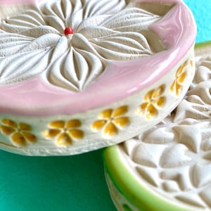 Könnte beinhalten: Zwei runde Keramikuntersetzer mit geprägten Blumenmustern. Der obere Untersetzer hat einen rosa glasierten Rand und einen roten Punkt in der Mitte. Die Seite ist mit gelben Blumenabdrücken versehen. Der untere Untersetzer hat einen grünen Rand.