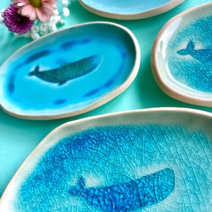Può includere: Quattro piattini ovali in ceramica con bordo chiaro e interno blu lucido. Tre dei piattini presentano una sagoma di balena blu scuro al centro, mentre uno ha una smaltatura blu chiaro uniforme. Le superfici presentano una finitura craquelé.