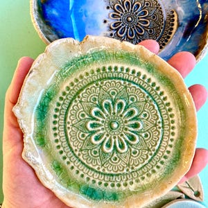 Op de afbeelding: Een handgemaakte keramische schaal met een groen mandala-ontwerp. De schaal heeft een getextureerd, gecraqueleerd oppervlak met een lichtgroen en beige kleurenschema. Een blauwe en bruine schaal staat op de achtergrond. Decoratieve keramische kunst.