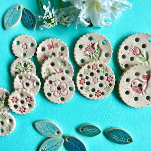 Può includere: Una collezione di sottobicchieri e ornamenti a forma di foglia in ceramica. I sottobicchieri sono bianco sporco con disegni floreali e di insetti in rosa e verde. Le foglie sono blu e verdi. Disposti su una superficie turchese con fiori bianchi.