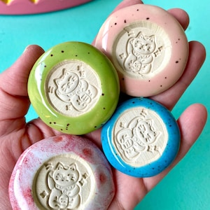 Könnte beinhalten: Vier kreisförmige Keramikplättchen mit einem eingeprägten Maneki-neko-Katzenmotiv in der Mitte. Die Plättchen sind in glänzendem Limettengrün, zartem Rosa, leuchtendem Blau sowie marmoriertem Weiß und Rot glasiert, jeweils mit kleinen dunklen Sprenkeln.