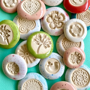 Peut inclure: Une collection de boutons en céramique ronds présentant divers motifs en relief, notamment des fleurs, des cœurs, des mandalas et des motifs botaniques. Les boutons ont des surfaces lisses et émaillées dans des tons rose, vert, bleu, rouge et blanc moucheté.