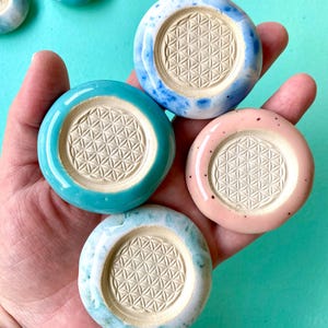 Può includere: Quattro gettoni circolari in ceramica con una finitura liscia e lucida nei toni del turchese, bianco maculato di blu e rosa tenue. Ogni gettone presenta un'indentazione circolare centrale non smaltata con un motivo geometrico a fiore.