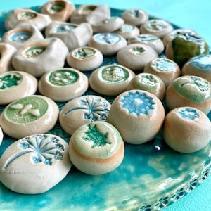 Puede incluir: Una colección de pequeñas piedras de cerámica con varios diseños en relieve en tonos verdes y azules. Las piedras están dispuestas sobre un plato de cerámica turquesa, creando una exhibición visualmente atractiva. Los diseños incluyen patrones florales y abstractos.