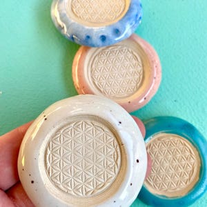 Pode incluir: Quatro bases para copos de cerâmica nas cores branca, azul claro, rosa e turquesa. Cada base apresenta um padrão floral em relevo no centro. As bases são redondas com uma borda ligeiramente elevada. Diâmetro de aproximadamente 10 cm.