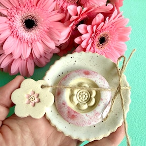 Könnte beinhalten: Eine runde, cremeweiße Keramikschale mit gesprenkelten Details und gewelltem Rand, die in der Mitte ein kreisförmiges Element mit rosafarbener Glasur und einem geformten Blumenmuster aufweist. Ein kleiner blumenförmiger Anhänger ist befestigt.