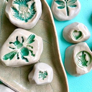 Puede incluir: Una colección de pequeñas piedras de cerámica blanquecina con diseños verdes en relieve. Los diseños incluyen un cardo, una libélula, una abeja, un corazón, una rana y una hoja. Las piedras están dispuestas sobre una bandeja de cerámica con forma de hoja.