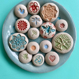 Puede incluir: Una colección de pequeñas piezas de arte cerámico redondas con varios diseños en relieve, incluyendo flores, una libélula, un pájaro y un árbol. Las piezas son en tonos de blanco, azul, rojo y verde, dispuestas sobre un plato de cerámica azul claro.