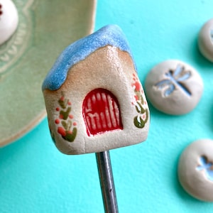 Pode incluir: Espeto de cerâmica em forma de casa com porta vermelha e telhado azul. A casa é decorada com detalhes florais verdes e vermelhos. Outras peças de cerâmica com desenhos de libélulas e flores estão no fundo.