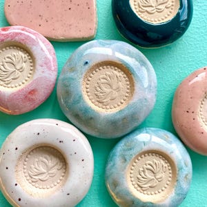 Könnte beinhalten: Eine Sammlung kreisförmiger Keramikobjekte mit glänzender Oberfläche in Rosa, Blau, Weiß und Dunkelgrün. Jedes Stück hat eine zentrale, unglasierte kreisförmige Vertiefung mit einem eingeprägten Lotusblumenmotiv und Perlenrand.