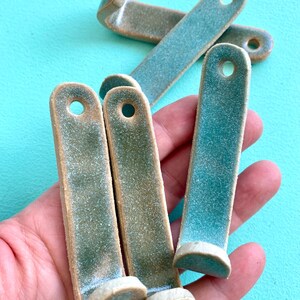 May include: A collection of handmade ceramic hooks in shades of teal and olive green. Each hook has a rounded end and a hole for hanging. The hooks are displayed against a bright turquoise background.