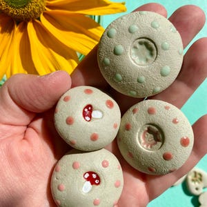 Puede incluir: Cuatro piedras decorativas redondas de cerámica beige con temática de setas, con lunares rojos y rosas y un diseño de seta roja. Dos piedras tienen una hendidura central. Un girasol amarillo está en el fondo.