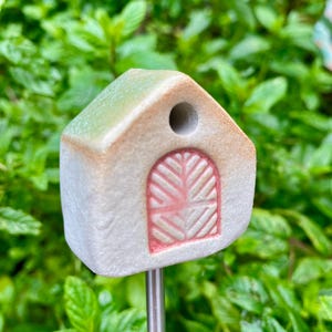 Peut inclure: Un petit piquet de jardin en céramique en forme de maison, avec une porte rose et un trou. Le toit est vert clair, monté sur une tige métallique argentée. Sur un fond de feuillage vert flou.
