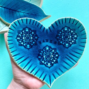 Op de afbeelding: Een hartvormige keramische schaal met een levendige blauwe glazuur en ingewikkelde mandala-ontwerpen. De schaal heeft een getextureerd oppervlak en is geaccentueerd met donkerdere blauwe lijnen. Twee bladvormige schalen staan op de achtergrond.