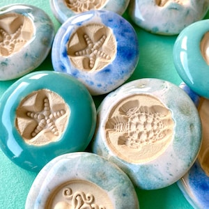 Könnte beinhalten: Eine Sammlung runder Keramik-Token mit geprägten Meeresmotiven wie Seesternen, einer Meeresschildkröte und einem Oktopus. Jeder Token hat einen glatten, glänzend glasierten Rand in Blau-, Türkis- und Weißtönen, der ein mattes, beiges Zentrum umgibt.