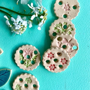 Könnte beinhalten: Eine Sammlung beigefarbener Keramikuntersetzer mit floralen und botanischen Designs in Rosa und Grün. Jeder Untersetzer hat einen gewellten Rand und drei Löcher. Weiße Blumen und blau-grüne Blätter im Hintergrund.