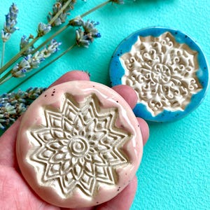 Può includere: Due dischi in ceramica rotondi con intricati motivi in rilievo in stile mandala. Un disco è rosa pallido lucido con sottili macchioline scure, mentre l'altro è turchese vibrante con macchioline simili. Entrambi hanno una finitura smaltata.