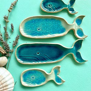 Può includere: Quattro piatti in ceramica a forma di balena di varie dimensioni, ciascuno caratterizzato da una finitura smaltata craquelé lucida nei toni del blu e del verde acqua. I piatti presentano un bordo in argilla chiara e dettagli incisi.