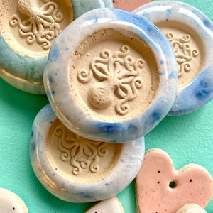 Könnte beinhalten: Eine Sammlung runder Keramikornamente mit einem erhabenen Oktopus-Relief in der Mitte, veredelt mit einer blau-weißen, gesprenkelten Glasur. Dazwischen befinden sich kleinere herzförmige Keramikstücke mit einer rosa-weißen, gepunkteten Oberfläche.