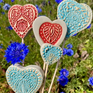 Peut inclure: Plusieurs tuteurs de jardin en céramique en forme de cœur, rouges et turquoise, avec des motifs en relief. Les tuteurs sont sur des bâtons en bois, sur fond de fleurs bleues et de verdure.