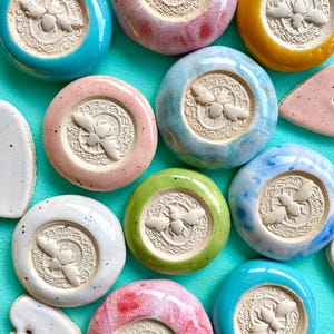 Op de afbeelding: Een verzameling kleurrijke keramische onderzetters met een bijenmotief. Elke onderzetter heeft een verhoogd bijenontwerp in een cirkelvormig frame. De onderzetters zijn geglazuurd in verschillende kleuren, waaronder blauw, roze, groen en geel. Sommige hebben gespikkelde details.