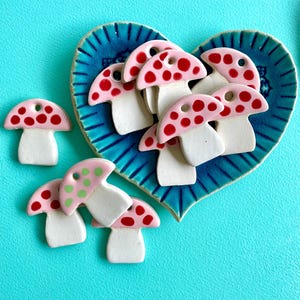 Könnte beinhalten: Keramik-Pilz-Ornamente mit rosa Kappen und roten und grünen Punkten. Sie sind auf einem herzförmigen blauen Keramikteller vor türkisfarbenem Hintergrund angeordnet. Dekorative Elemente.