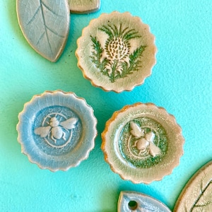 Puede incluir: Platos decorativos de cerámica y adornos en forma de hoja en tonos azul, verde y crema. Los platos presentan diseños en relieve de una abeja y un cardo. Los adornos de hojas tienen un agujero para colgar.