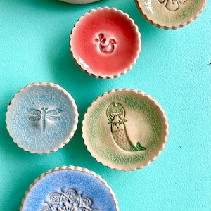 Puede incluir: Una colección de pequeños platos decorativos de cerámica en varios colores, incluyendo verde, rosa, azul y beige. Cada plato presenta un diseño en relieve único, como una libélula, una flor, una sirena o un mandala. Los platos tienen un borde festoneado.