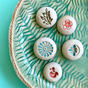 Puede incluir: Pequeñas piedras redondas de cerámica blanquecina con diseños en relieve, incluyendo una flor, una ramita y un pájaro. Están dispuestas sobre un plato de cerámica verde con un patrón de hojas de helecho.