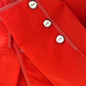 May include: Red fabric with white stitching and three silver buttons. The fabric is folded over itself.