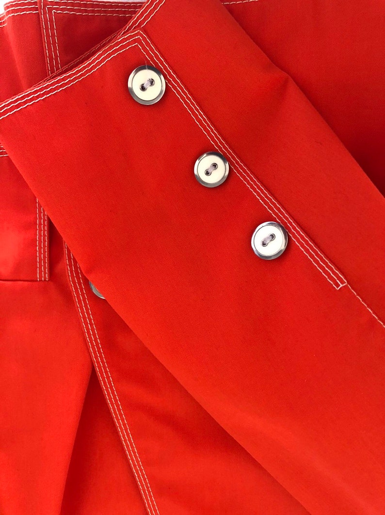 May include: Red fabric with white stitching and three silver buttons. The fabric is folded over itself.