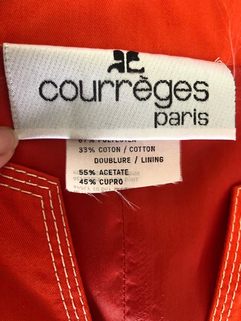May include: A white tag with black text that reads "courr&egrave;ges paris". The tag is attached to a red fabric garment. The tag also includes the following information: 67% Polyester, 33% Cotton, Doublure/Lining 55% Acetate, 45% Cupro.