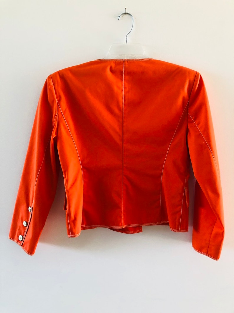 May include: A bright orange blazer with long sleeves and white stitching. The blazer has a single button on each cuff and a single button on the back.
