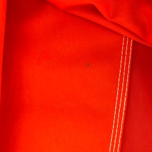 May include: Close-up of a bright orange fabric with white stitching. The fabric has a subtle texture and a small brown stain.